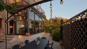Un área de comedor exterior moderna con un porche acristalado, luces colgantes y vistas al interior.