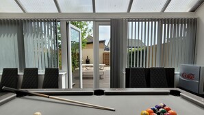 A pool table with a cue stick and balls, a Diet Coke cooler, and a view of a backyard through large windows. - House of 4 Chimneys by Your Lettings UK (Peterborough)