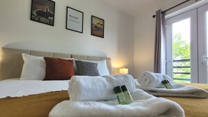 A hotel room with a neatly made bed, two rolled towels, and a bottle of lotion on the bed. - House of 4 Chimneys by Your Lettings UK (Peterborough)