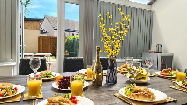 A breakfast table with plates of food, glasses of orange juice, and a bottle of champagne. - House of 4 Chimneys by Your Lettings UK (Peterborough)