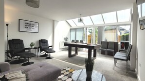 A modern living room with a sofa, chairs, a coffee table, and a dining table. There is a large window with blinds, a ceiling fan, and a framed picture on the wall. - House of 4 Chimneys by Your Lettings UK (Peterborough)