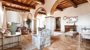 A spacious living room with a high wooden ceiling, arched brick columns, and a large window. - I Tebaldi - Eight Bedroom Villa, Sleeps 18 (Reggello)