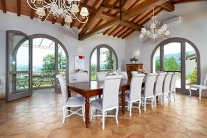 A dining area with a large wooden table and white chairs, large arched windows, and a view of greenery outside. - I Tebaldi - Eight Bedroom Villa, Sleeps 18 (Reggello)