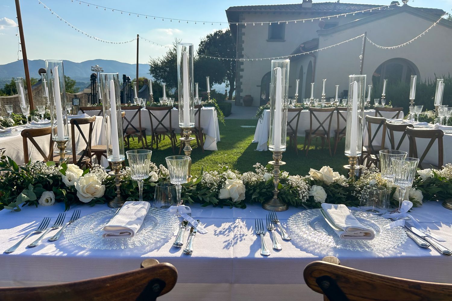 An outdoor dining area with tables set for a formal event, featuring clear glassware, white tablecloths, and floral centerpieces.