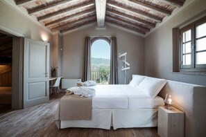 A bedroom with a bed, a bedside table with a lamp, a window with a view, and a door leading to another room. - I Tebaldi - Eight Bedroom Villa, Sleeps 18 (Reggello)