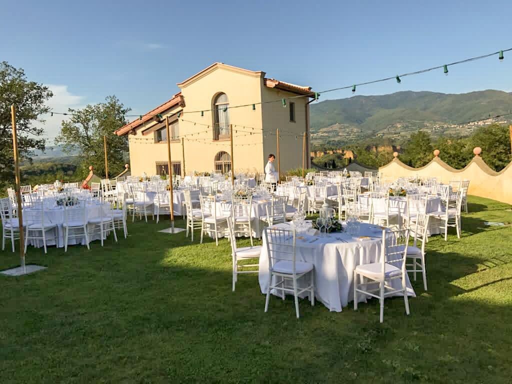 An outdoor wedding venue with multiple round tables and chairs, a building in the background, and a view of hills.