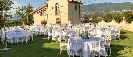 An outdoor wedding venue with multiple round tables and chairs, a building in the background, and a view of hills.