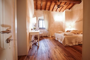 A room with a wooden floor, a bed with a white bedspread, a desk with a lamp, and a window with curtains. - I Tebaldi - Eight Bedroom Villa, Sleeps 18 (Reggello)