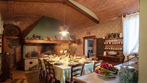 A rustic dining room with a brick ceiling, wooden tables, and chairs. There is a brick fireplace, shelves with decorative items, and a window with curtains. - Tenuta Bartolucci - Ten Bedroom Villa, Sleeps 22 (Palaia)