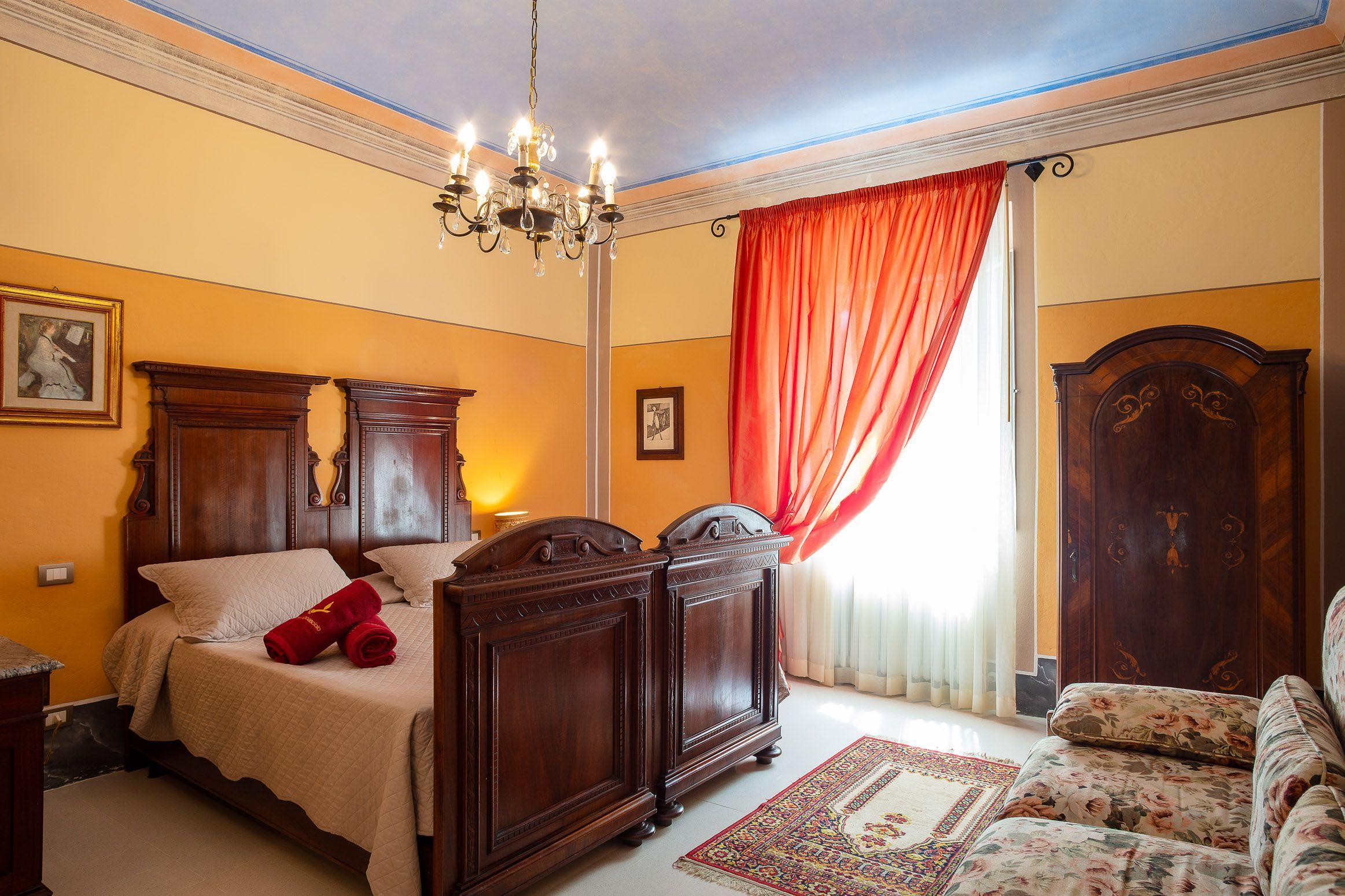A hotel room with a large bed, a wooden wardrobe, a chandelier, and a floral sofa.
