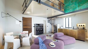 Un espace de vie moderne comprenant une table à manger en verre, des chaises blanches, un canapé violet, un coin cuisine et un escalier menant à un niveau supérieur.