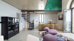 Un salon moderne avec une table basse en verre, des canapés violets, un meuble noir avec des portes en verre, un escalier en colimaçon et un coin cuisine avec un bar.