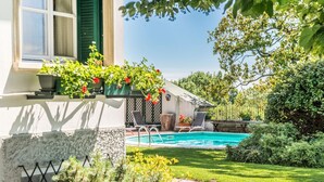 Un área junto a la piscina con un muro de piedra, un pequeño jardín y un edificio con una ventana y persianas verdes.