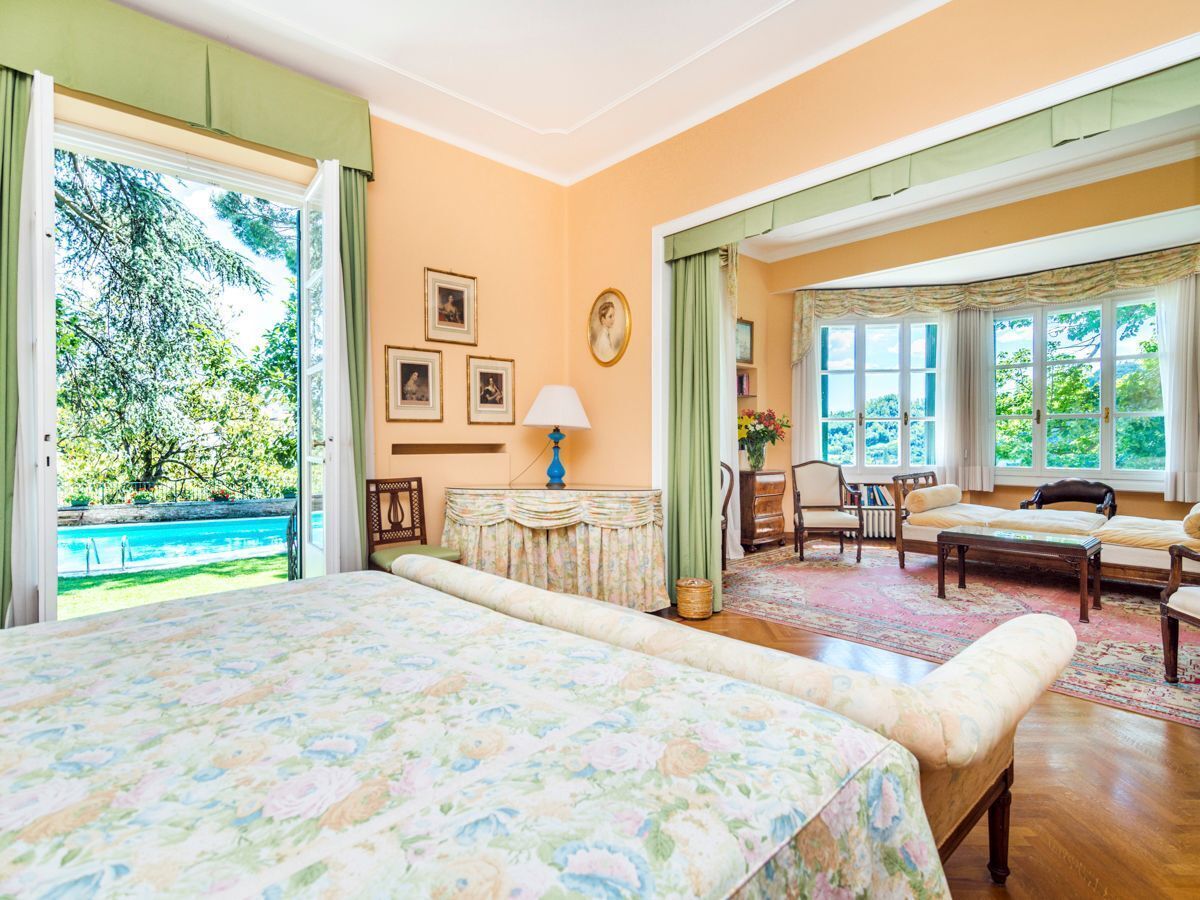 Un'ampia camera da letto con un grande letto, un'area salotto con divano e poltrone, e vista su una piscina e vegetazione.