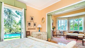 Un dormitorio amplio con una cama grande, un área de estar con sofá y sillones, y vistas a una piscina y zonas verdes.