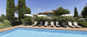 A swimming pool with lounge chairs and umbrellas, surrounded by greenery and a building in the background.