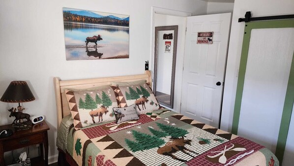 Un dormitorio con una cama que presenta ropa de cama con motivos de alce y bosque, un cuadro de un alce junto a un lago, una puerta con una señal de advertencia de alce y una mesita de noche con una lámpara de alce.