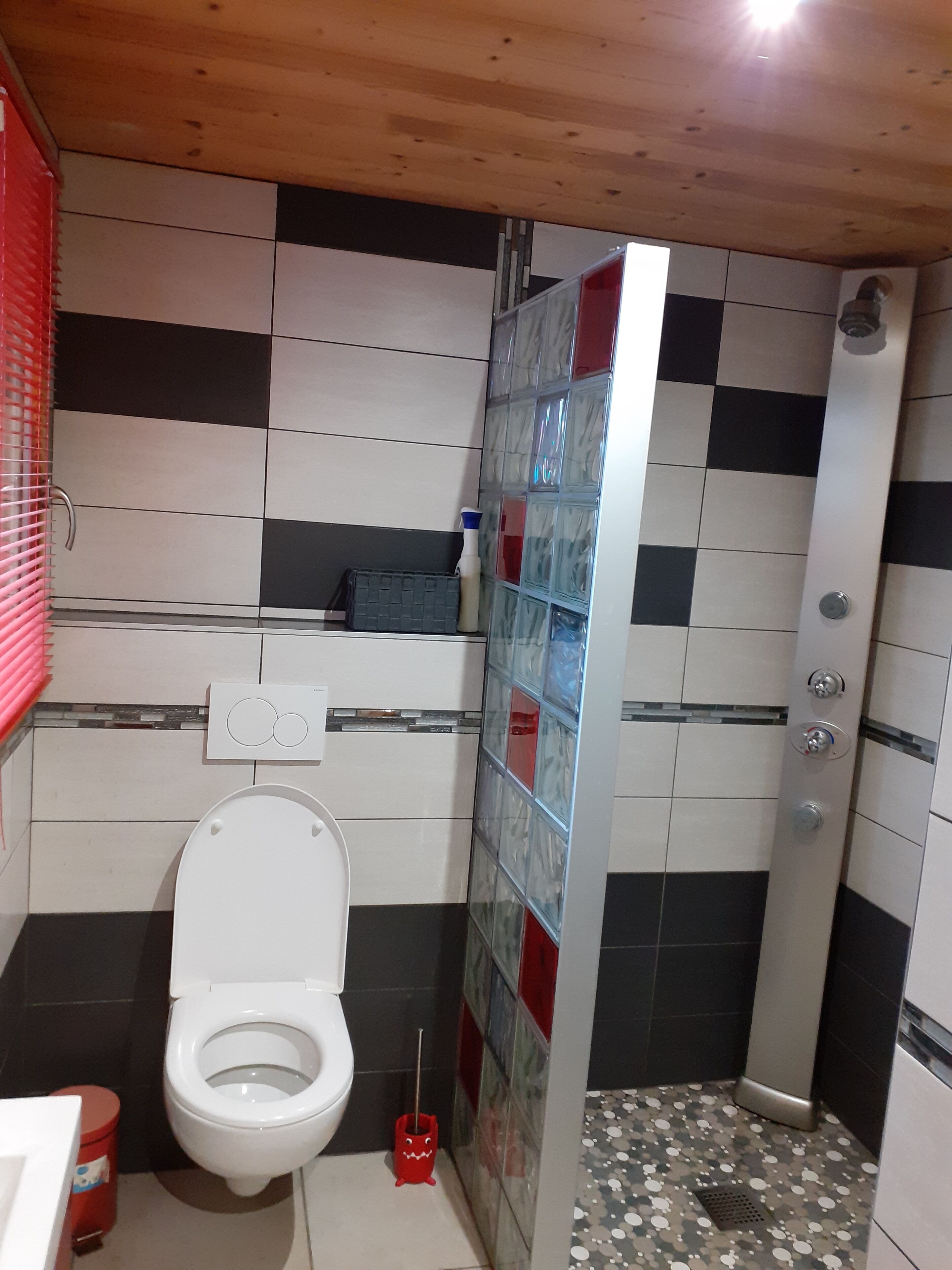 Un baño con inodoro blanco, un cubo rojo y una zona de ducha con paredes de azulejos.