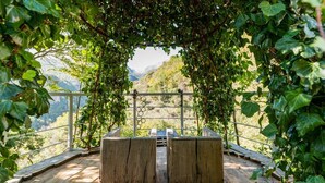 Une terrasse en bois avec deux bancs, située sous une pergola couverte de plantes grimpantes, et offrant une vue sur un paysage montagneux.