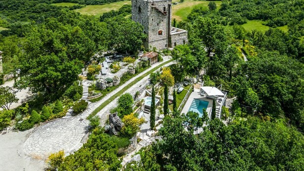 Une vue aérienne d’un château entouré d’une végétation luxuriante, avec une tour en pierre, une piscine et une cour intérieure circulaire en pierre.
