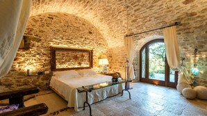 Une chambre aux murs de pierre, avec un lit, une table de chevet, une lampe et une vue sur l’extérieur par une fenêtre en arc de cercle.