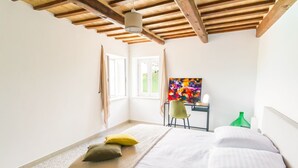 Een slaapkamer met een bed, een bureau en een stoel. Er zijn ramen met gordijnen, een schilderij aan de muur en een groene fles op het nachtkastje.