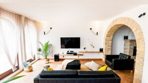 Een moderne woonkamer met een zwarte bank, een flatscreen televisie, een open haard en een groot raam met dunne gordijnen.