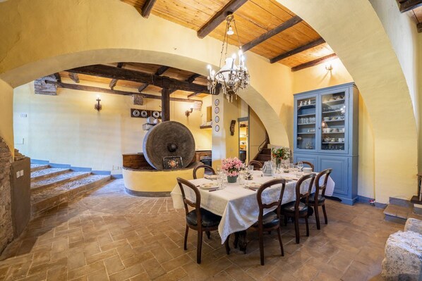 A dining area with a round table set for six, surrounded by chairs. A chandelier hangs from the ceiling, and a blue cabinet with glassware is visible in the background.