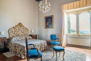 Un dormitorio con una cama grande, dos sillones tapizados en azul, una araña, un cuadro en la pared y una ventana con vistas.
