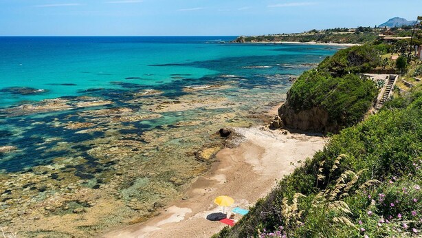 Un paysage côtier caractérisé par une eau turquoise cristalline, des rivages rocheux et un sentier menant à une plage parsemée de parasols.