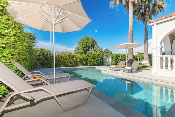 A swimming pool with lounge chairs and umbrellas, surrounded by greenery and a white building.