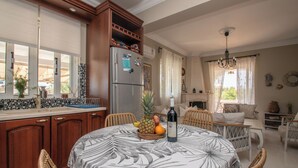 Une cuisine avec des meubles en bois, un réfrigérateur, une table à manger recouverte d’une nappe à motifs tropicaux et un panier de fruits.