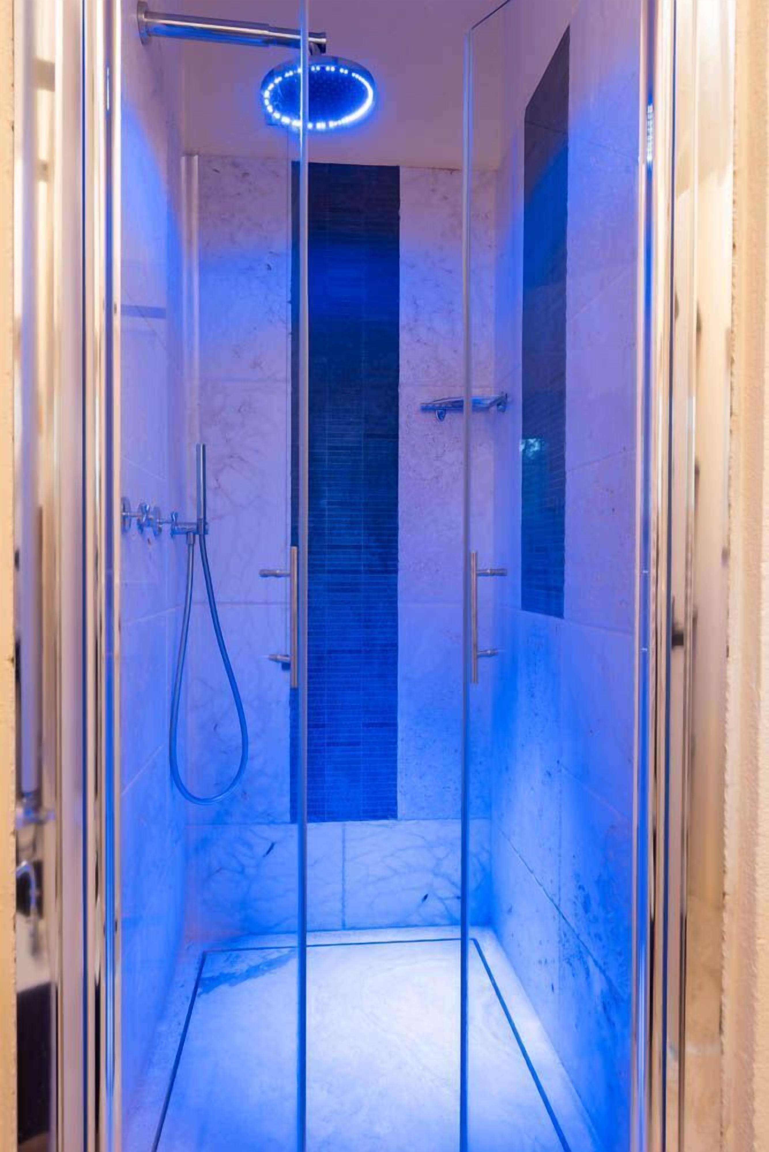 Ducha con paredes de vidrio, una lámpara azul y un cabezal de ducha manual, además de una puerta de vidrio vertical.