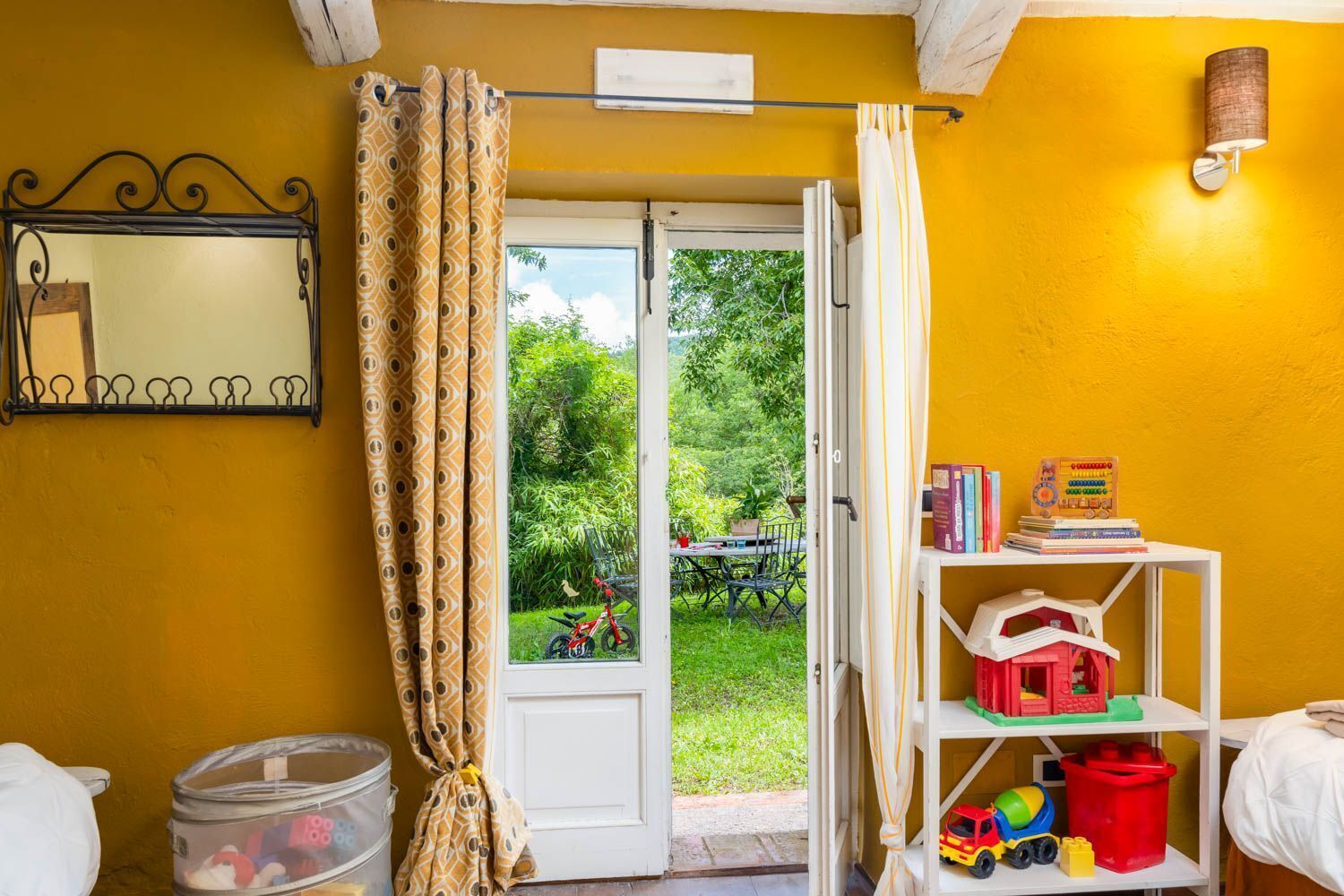 A room with a yellow wall, a white door leading to a garden, a metal rack with keys, a shelf with children's toys, and a small table with books.