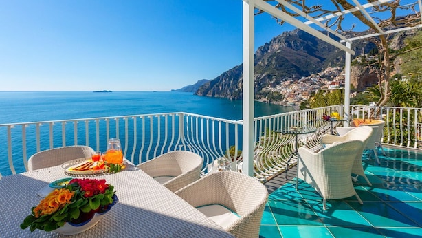 Eine Terrasse mit Korbmöbeln, einem Tisch mit Getränken und Blumen sowie Blick auf das Meer und die Berge.