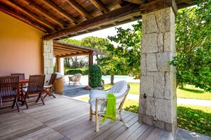 An outdoor patio with wooden flooring, a stone pillar, and a covered seating area with chairs and a table.