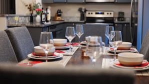 A dining table set with plates, glasses, and cutlery, with a kitchen in the background.