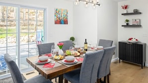 A dining area with a wooden table set for four, grey chairs, a chandelier, and a view through a glass door to an outdoor area.