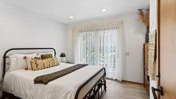 A bedroom with a bed, bedside table, lamp, and a view of the outdoors through a window.