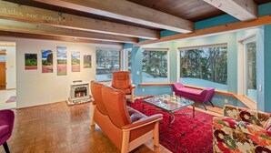 Un salon chaleureux avec une cheminée, des poutres en bois, de grandes fenêtres et une moquette rouge.