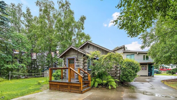 Une cabane en rondins dotée d’une terrasse en bois et d’un petit porche, entourée d’une végétation luxuriante et bordée d’une allée pavée.
