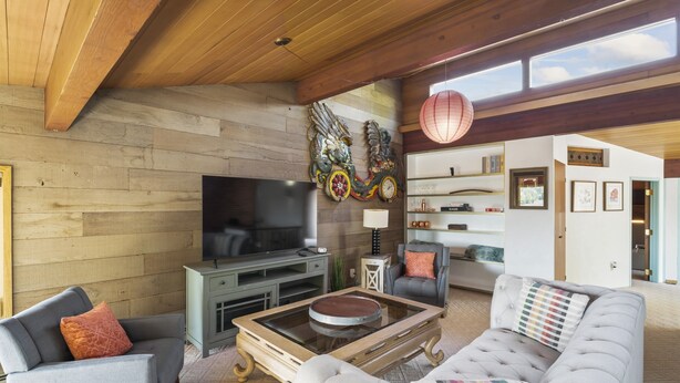 A modern living room with a wooden ceiling, a flat-screen TV, a glass coffee table, and a grey sofa.