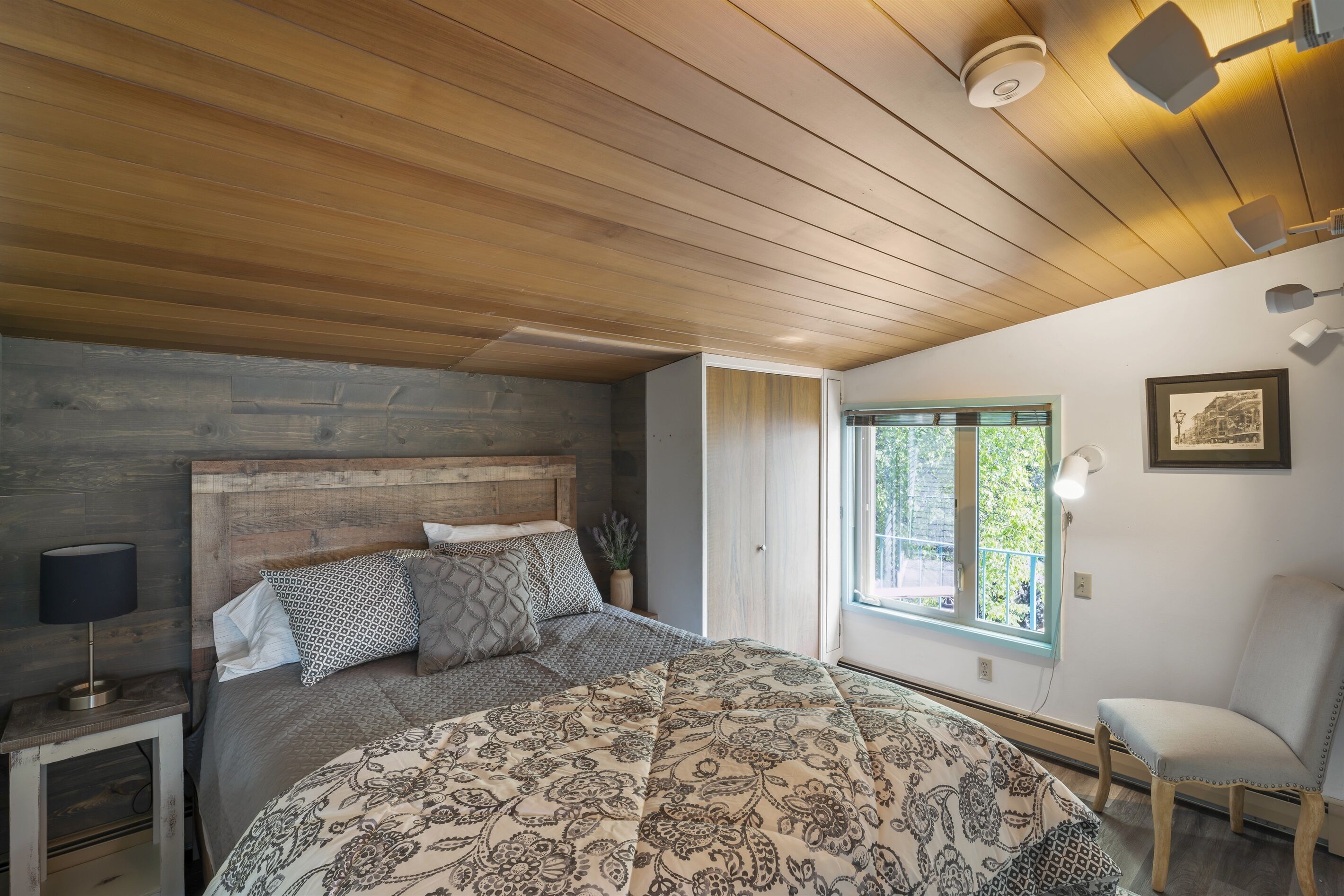 A bedroom with a wooden ceiling, a bed with patterned bedding, a nightstand with a lamp, a chair, and a window with a view of greenery.