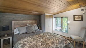 A bedroom with a wooden ceiling, a bed with patterned bedding, a nightstand with a lamp, a chair, and a window with a view of greenery.