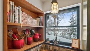 A kitchen with a wooden shelf holding books, kitchen utensils, and red mugs. A window with a view of a snowy landscape is visible. - Historic, Heart of City Home - Near U/Med District (Anchorage)