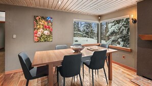 A dining area with a wooden table and four chairs, a large floral artwork on the wall, and a window with a snowy landscape view. - Historic, Heart of City Home - Near U/Med District (Anchorage)