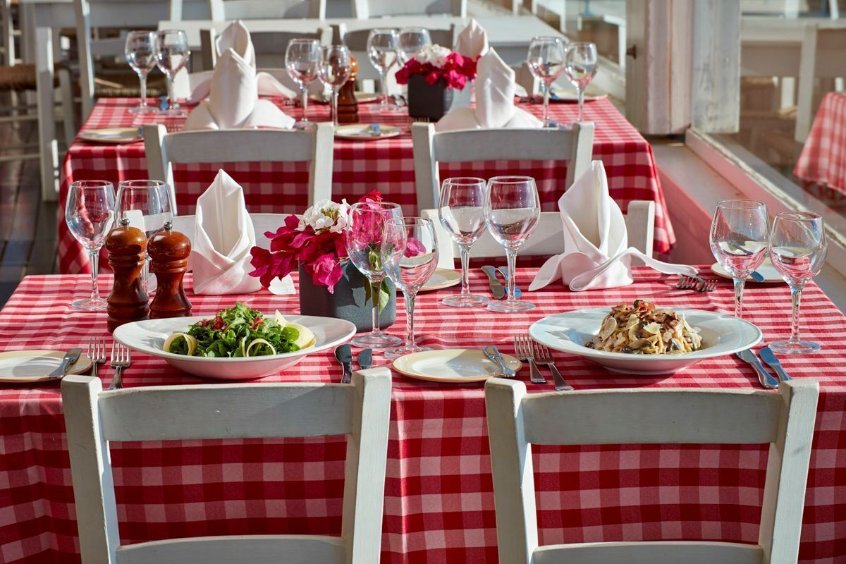 Un restaurante con manteles a cuadros rojos y blancos, sillas blancas y mesas de comedor ordenadas, con platos de comida, vasos y cubiertos.