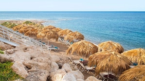 A beach with thatched umbrellas and lounge chairs, a rocky shoreline, and the sea in the background.
