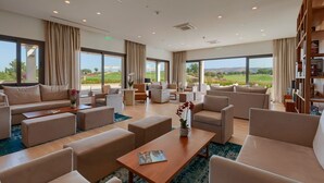 A spacious, well-lit lounge area with multiple seating options, large windows offering a view of the outdoors, and a bookshelf filled with books.