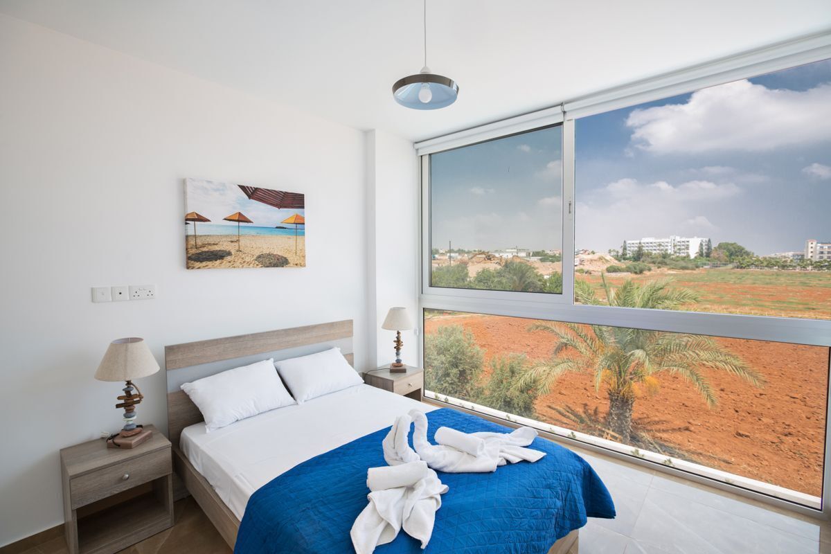 Een slaapkamer met een bed, nachtkastje, lamp, schilderij van een strandgezicht en een groot raam met uitzicht op een landschap.